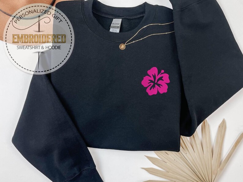 Puede incluir: Sudadera negra con un dise&ntilde;o de flor de hibisco rosa. La sudadera tiene mangas largas y cuello redondo. Un collar de oro con un peque&ntilde;o colgante est&aacute; drapeado sobre la prenda. El texto "REGALO PERSONALIZADO SUDADERA Y SUDADERA CON CAPUCHA BORDADA" est&aacute; en un dise&ntilde;o circular.