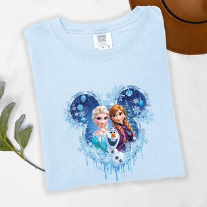 May include: Light blue Comfort Colors t-shirt featuring a graphic of Elsa, Anna, and Olaf from Disney's Frozen. The design is set within a Mickey Mouse head shape with snowflakes and icicles. The shirt has a crew neck and short sleeves.