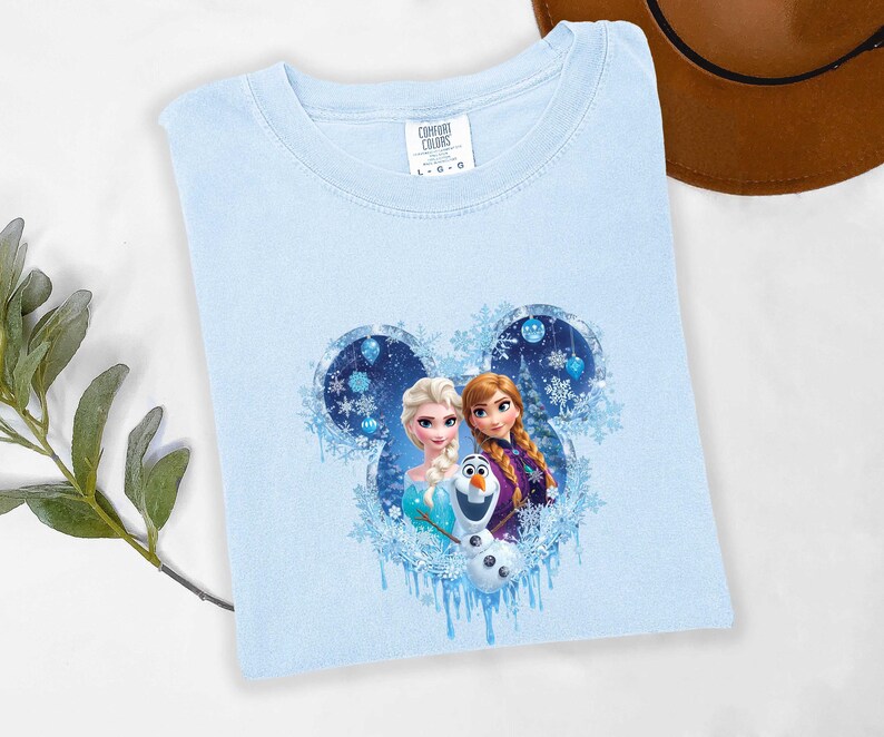 May include: Light blue Comfort Colors t-shirt featuring a graphic of Elsa, Anna, and Olaf from Disney's Frozen. The design is set within a Mickey Mouse head shape with snowflakes and icicles. The shirt has a crew neck and short sleeves.