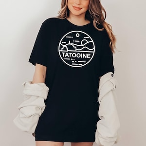 May include: Black t-shirt with a white circular graphic featuring the word "TATOOINE" and a landscape design. The shirt is a crew neck style and appears to be made of a soft material.