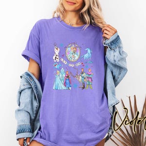 May include: A purple t-shirt featuring a colorful graphic of characters from the movie "Frozen", including Olaf, Elsa, Anna, Kristoff, and Sven. The phrase "Let It Go" is also displayed. The shirt is a comfortable, oversized fit.