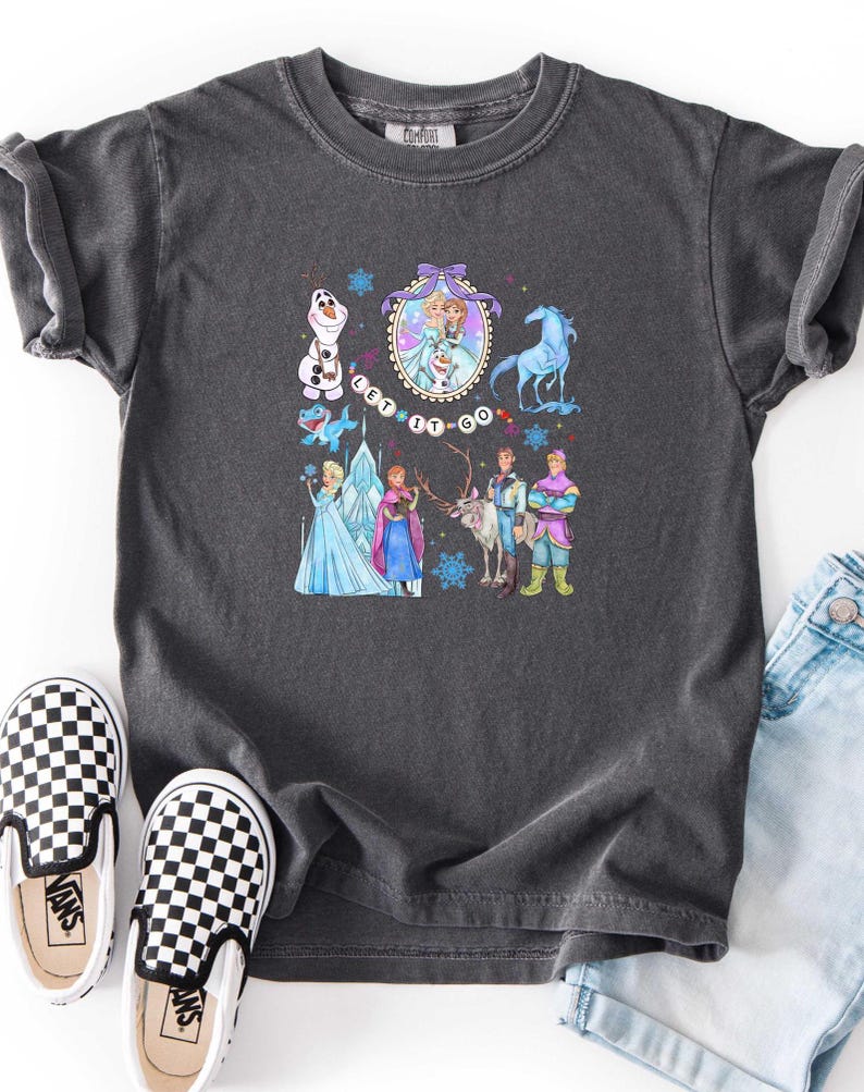 May include: Dark gray t-shirt featuring a colorful graphic of characters from the movie "Frozen", including Elsa, Anna, Olaf, and Kristoff. The phrase "Let It Go" is also displayed. The shirt is paired with black and white checkered slip-on shoes.