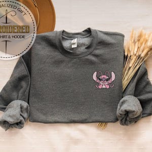 Puede incluir: Una sudadera gris oscuro bordada con un dise&ntilde;o de personaje de dibujos animados rosa. La sudadera tiene mangas largas y cuello redondo. La imagen tambi&eacute;n incluye el texto "PERSONALIZED GIFT EMBROIDERED SWEATSHIRT & HOODIE".