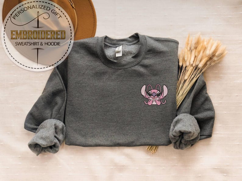 Puede incluir: Una sudadera gris oscuro bordada con un dise&ntilde;o de personaje de dibujos animados rosa. La sudadera tiene mangas largas y cuello redondo. La imagen tambi&eacute;n incluye el texto "PERSONALIZED GIFT EMBROIDERED SWEATSHIRT & HOODIE".