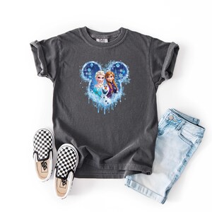 May include: Dark gray t-shirt featuring a Disney Frozen graphic with Elsa, Anna, and Olaf. The shirt is paired with light blue denim shorts and black and white checkered slip-on shoes.