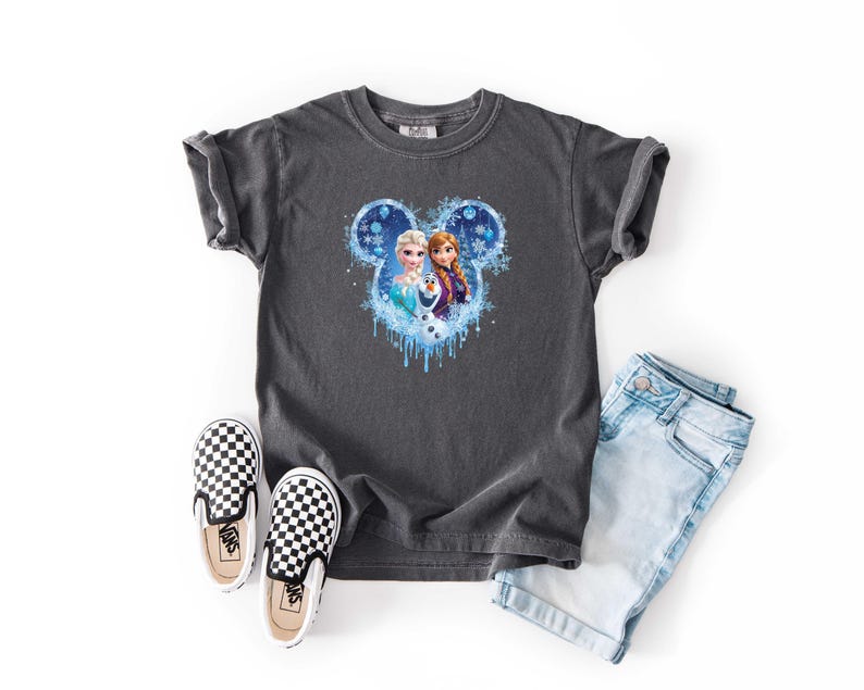 May include: Dark gray t-shirt featuring a Disney Frozen graphic with Elsa, Anna, and Olaf. The shirt is paired with light blue denim shorts and black and white checkered slip-on shoes.