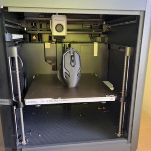 May include: A black 3D printer with a black mouse on the print bed. The printer has a clear plastic door and a small screen on the top. The print bed is labeled "256 x 256 x 256 mm". The printer is labeled "Bambu Lab P1S".