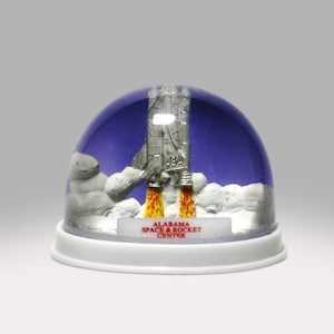 May include: A snow globe featuring a silver rocket launching against a blue backdrop. The rocket has orange flames and is set on a white base with the text "ALABAMA SPACE & ROCKET CENTER". The globe is encased in a clear dome.