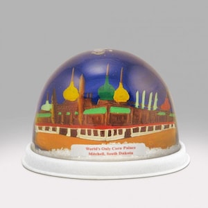 Könnte beinhalten: Eine Schneekugel mit dem Corn Palace in Mitchell, South Dakota. Die Kuppel enthält eine blaue Flüssigkeit und ein farbenfrohes Gebäude mit gelben, grünen und braunen Details. Der weiße Sockel trägt den Text "World's Only Corn Palace Mitchell, South Dakota".
