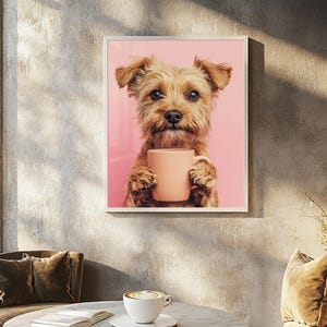 May include: A framed print featuring a Yorkshire Terrier dog holding a peach-colored mug against a pink background. The dog has brown fur and is looking directly at the viewer. The print is displayed on a textured gray wall.