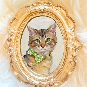 Op de afbeelding: Ovale schildering van een kat met een groene vlinderdas, omlijst in goud. De kat heeft bruin en grijs haar en groene ogen. De gouden lijst is sierlijk en in vintage stijl, met decoratieve details.