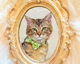 May include: An oval-shaped painting of a cat wearing a green bow tie, set within a gold ornate frame. The cat has brown and gray fur with green eyes. The frame is a detailed, vintage style with decorative flourishes.