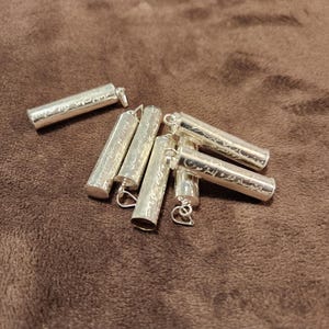 Puede incluir: Un grupo de seis colgantes cilíndricos de plata con intrincados diseños grabados. Los colgantes tienen un bucle en la parte superior para sujetarlos a una cadena o cordón.