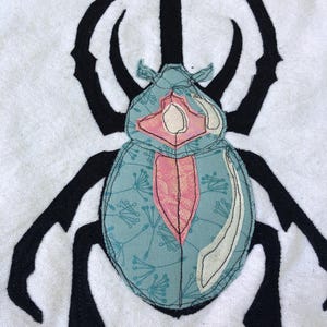 May include: A white fabric patch featuring a black silhouette of a beetle. The beetle has a light blue body with a delicate floral pattern, accented by a central pink section and a white curved detail, all outlined with visible black stitching.