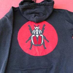 May include: A black hooded sweatshirt featuring a large circular red patch on the back. Centered on the red circle is a stylised black and white beetle graphic with two prominent red textured wing covers. The hoodie includes a drawstring hood.