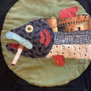 May include: A black hoodie featuring a large circular olive green fabric patch. The patch displays a fish appliqué with a dark patterned head, a cigarette in its mouth, and a body section depicting a stone castle wall with red fins.
