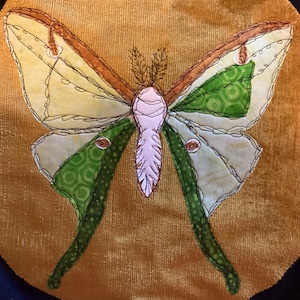May include: A textile applique of a moth with a white body and feathery antennae, featuring wings in shades of light yellow, lime green, and dark green with brown stitched outlines, set against a textured, mustard-yellow fabric background.