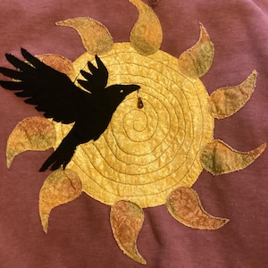 May include: A mauve sweatshirt featuring a large, circular yellow sun appliqué with a spiral-stitched center and patterned rays. A black fabric crow is appliquéd over the sun, holding a small, dangling brown bead in its beak.