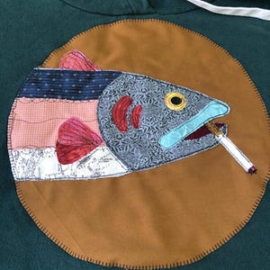 May include: A dark green hooded sweatshirt featuring a large circular brown fabric patch appliqued on the front. The patch displays a multi-patterned fabric fish with red fins, a grey floral head, and a cigarette held in its mouth.