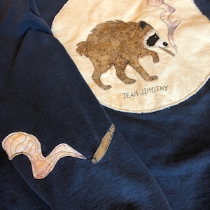 May include: A navy blue sweatshirt featuring a large, circular cream-colored fabric patch on the chest with an embroidered brown raccoon smoking a cigarette and the text "TEAM JIMOTHY". A separate patch of a cigarette and smoke is on the sleeve.