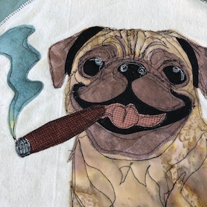May include: A circular fabric patch featuring a tan pug with a black muzzle and ears, holding a brown cigar in its mouth. The pug is depicted with a pink tongue and large dark eyes. A trail of blue smoke rises from the cigar against a white background.