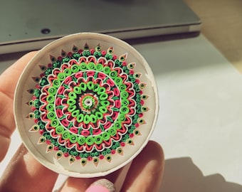 May include: A close-up of a circular mandala made of paper, with a white background and a design of green, pink, and red swirls.
