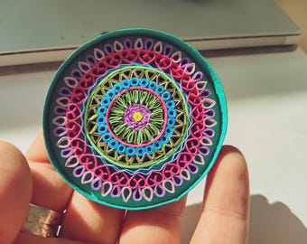 May include: A colorful paper quilling mandala with a teal background. The mandala features concentric circles of pink, purple, blue, green, and yellow paper.