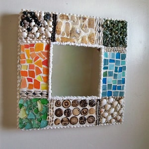 May include: Square mirror with a mosaic frame. The frame is decorated with a variety of materials, including shells, stones, and glass tiles in shades of orange, green, blue, and brown. The mirror is a decorative wall hanging.