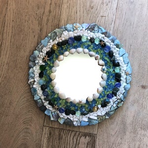 May include: A round mirror with a mosaic frame made of seashells, small stones, and glass tiles in shades of blue, green, and white. The mirror is surrounded by a border of white and blue seashells.