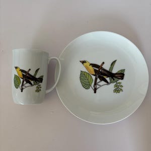 May include: White ceramic mug and plate set. Each piece features a detailed illustration of a yellow and black bird perched on a branch with green leaves. The mug has a handle, and the plate is round.