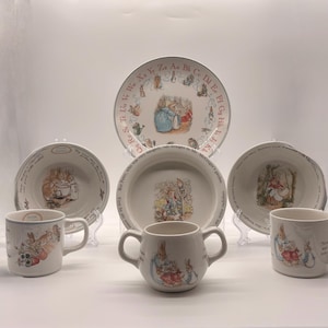May include: A collection of white ceramic tableware featuring illustrations of rabbits. The set includes plates, bowls, and mugs, with a light blue trim. The designs are reminiscent of children's storybook illustrations.