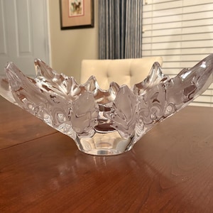 May include: A clear glass decorative bowl with a leaf design. The bowl has a wide, open shape with a series of leaf-like extensions. The bowl is sitting on a wooden table. The background includes a door, framed artwork, and window blinds.