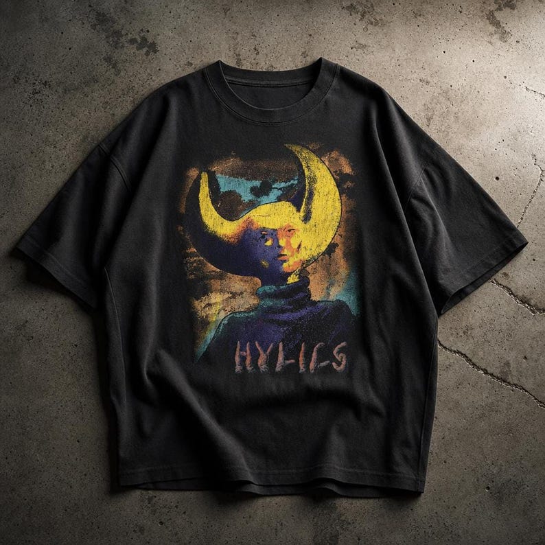 A black short-sleeved cotton t-shirt laid flat on a concrete surface, featuring a colorful graphic of a figure with large curved horns and the text Hylics.