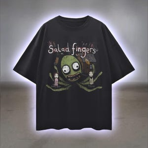 Puede incluir: Camiseta negra con la frase "Salad Fingers" sobre una ilustración de dibujos animados de una criatura verde con ojos grandes y una sonrisa amplia. El diseño incluye dos pequeñas figuras y una casa.
