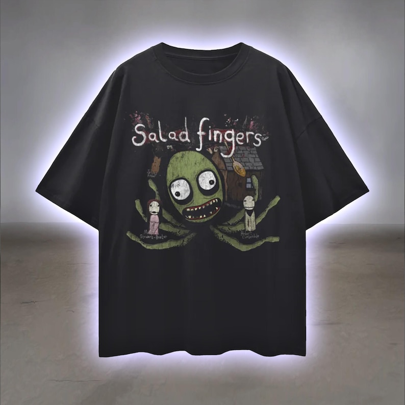 Peut inclure: T-shirt noir avec l'inscription "Salad Fingers" au-dessus d'un personnage de dessin anim&eacute; vert aux grands yeux et &agrave; la bouche souriante. Le motif comprend deux petits personnages et une maison.
