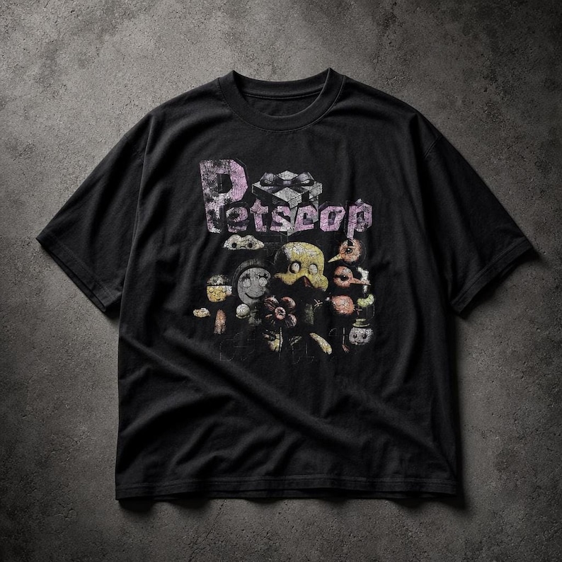 A black short-sleeve t-shirt flat on a textured grey surface, featuring a graphic print with the text Petscop and various quirky character illustrations.