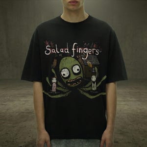 Peut inclure: T-shirt noir avec un dessin d'une cr&eacute;ature verte aux grands yeux et un large sourire, avec les mots "Salad Fingers" au-dessus. Le motif comprend une petite maison et deux personnages. Le t-shirt a des manches courtes.