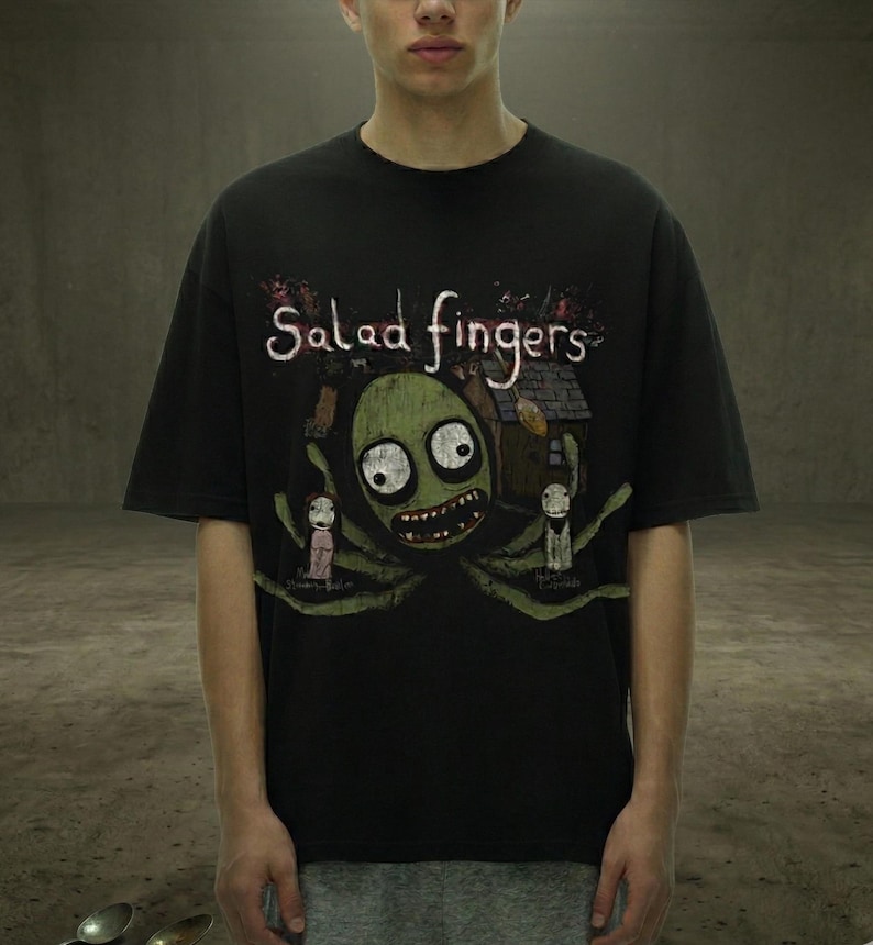 Peut inclure: T-shirt noir avec un dessin d'une cr&eacute;ature verte aux grands yeux et un large sourire, avec les mots "Salad Fingers" au-dessus. Le motif comprend une petite maison et deux personnages. Le t-shirt a des manches courtes.