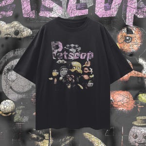 以下が含まれることがあります： 黒いTシャツ。テキスト「Petscop」は、使い古された紫色のフォントで表示されています。このシャツは、さまざまな色の漫画のようなキャラクターのグラフィックが特徴です。背景は、抽象的な形をした暗いテクスチャのデザインです。