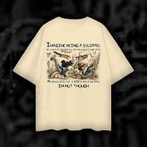 Puede incluir: Una camiseta beige con un estampado gráfico de ardillas en un entorno de bosque. El texto en la camiseta dice "Imagine being a squirrel. We could eat acorns all day. We could climb trees. We would be so tuff. I wish I was a squirrel. I'm not though."