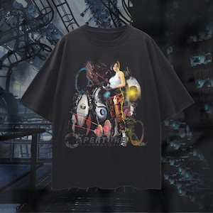 以下が含まれることがあります： ビデオゲームPortalのカラフルなグラフィックが特徴の黒いTシャツ。デザインには女性キャラクター、ロボット要素、そして「Aperture Laboratories」の文字が含まれています。このシャツは半袖で、クルーネックです。