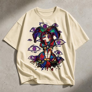 May include: A cream-coloured t-shirt with a vibrant graphic. The design depicts a jester-like character with a harlequin hat and outfit, surrounded by purple eyes. The artwork includes splashes of red, blue, and green, creating a striking visual.