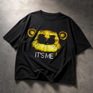 Op de afbeelding: Zwart T-shirt met een gouden berengezicht-afbeelding en de witte tekst "IT'S ME". Het gezicht van de beer heeft een verweerde, getextureerde uitstraling. Het shirt is gemaakt van een zacht materiaal en heeft korte mouwen.