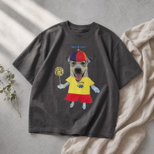 Könnte beinhalten: Dunkelgraues T-Shirt mit einem Hund, der einen Hut mit Propeller, ein gelbes Hemd und rote Shorts trägt. Der Hund hält einen bunten Lutscher. Das Shirt hat eine Grafik einer Zeichentrickfigur.