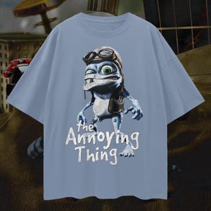 May include: A light blue t-shirt featuring a cartoon frog character wearing a pilot's hat and goggles. The text "the Annoying Thing" is printed in white below the character. The shirt has short sleeves and a crew neck.