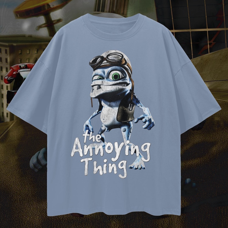 May include: A light blue t-shirt featuring a cartoon frog character wearing a pilot's hat and goggles. The text "the Annoying Thing" is printed in white below the character. The shirt has short sleeves and a crew neck.
