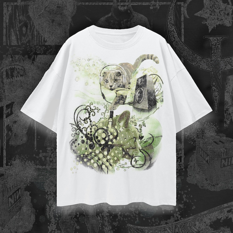 May include: A white short-sleeve t-shirt featuring a graphic print of two cats wearing headphones. The design includes a speaker, swirling black decorative lines, and green watercolor-style accents. The cats are positioned centrally on the chest.