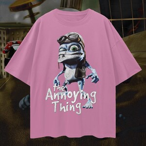 May include: Pink t-shirt featuring a cartoon frog character wearing a pilot's hat and goggles. The frog has blue skin and green eyes. The text "the Annoying Thing" is printed below the frog in white.