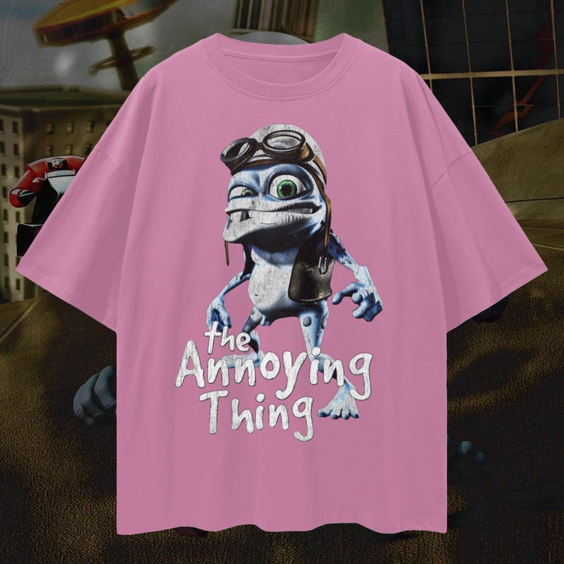 May include: Pink t-shirt featuring a cartoon frog character wearing a pilot's hat and goggles. The frog has blue skin and green eyes. The text "the Annoying Thing" is printed below the frog in white.