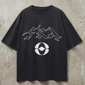 Può includere: T-shirt nero con testo grafico bianco e un design circolare. Il testo, "FKA", è stilizzato e posizionato sopra il design circolare. La maglietta ha maniche corte e girocollo. Lo sfondo è un muro neutro e strutturato.